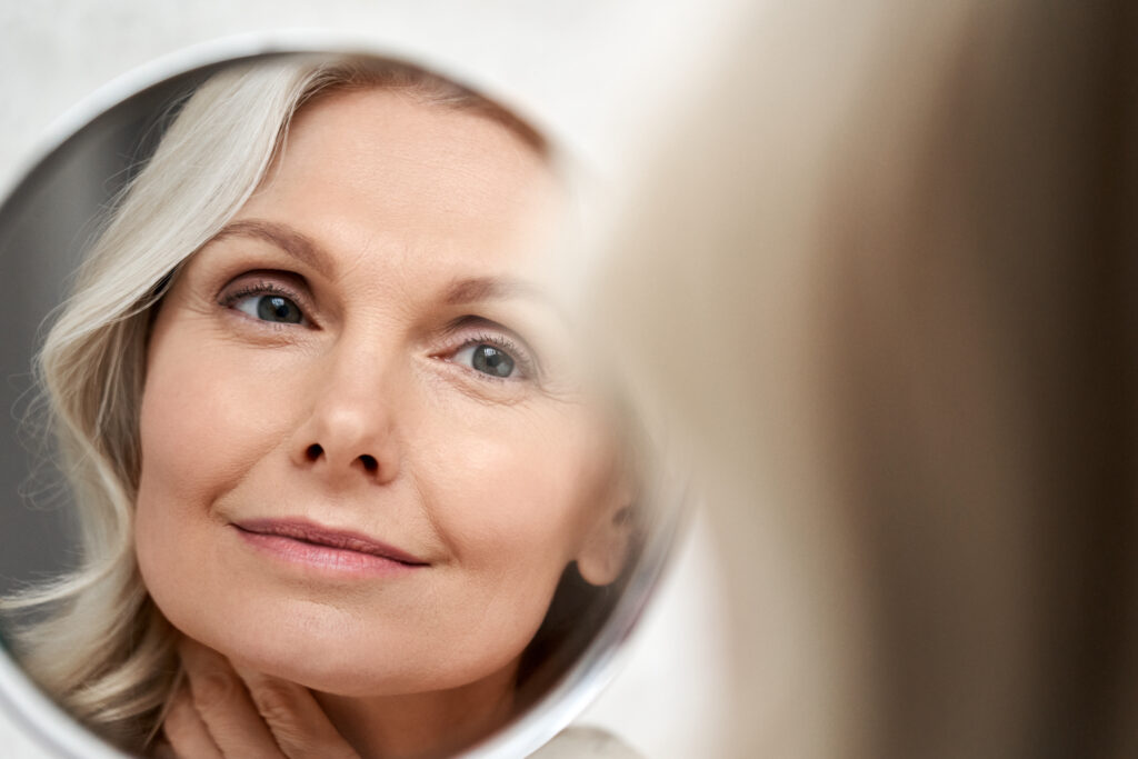 Happy 50s middle aged woman model touching face skin looking in mirror reflection.